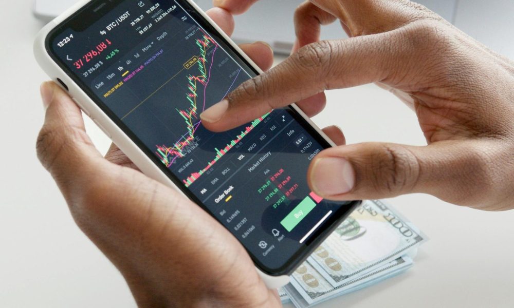 Hands using smartphone trading app with charts, dollar bills in background.